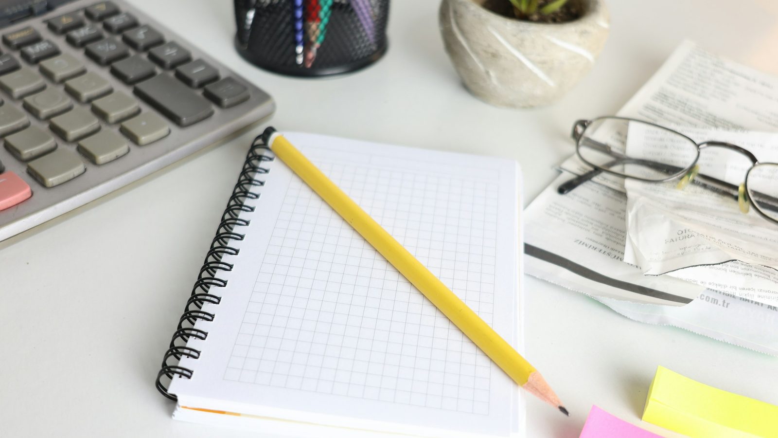 A notebook, calculator, pencil and reading glasses on a desk.